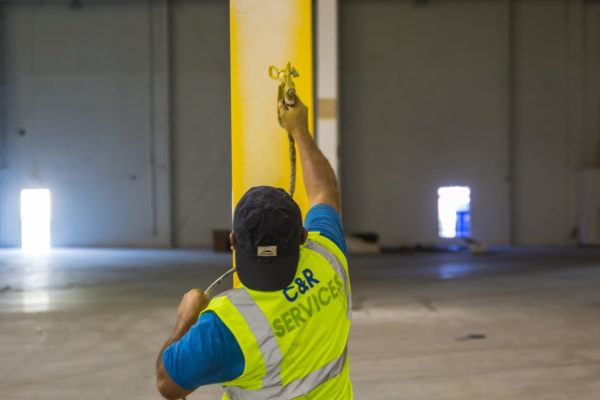 A man in a C&R Services vest airbrush paints a bright yellow column