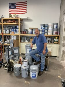 A man smiles as he poses, one foot on top of a stack of commercial paint cans.