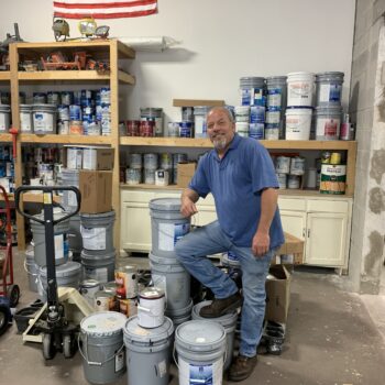 A man smiles as he poses, one foot on top of a stack of commercial paint cans.