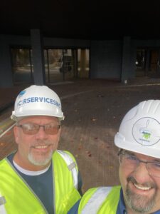 Two men in C&R Services hard hats and high-visibility yellow vests smile up at the camera in a selfie.