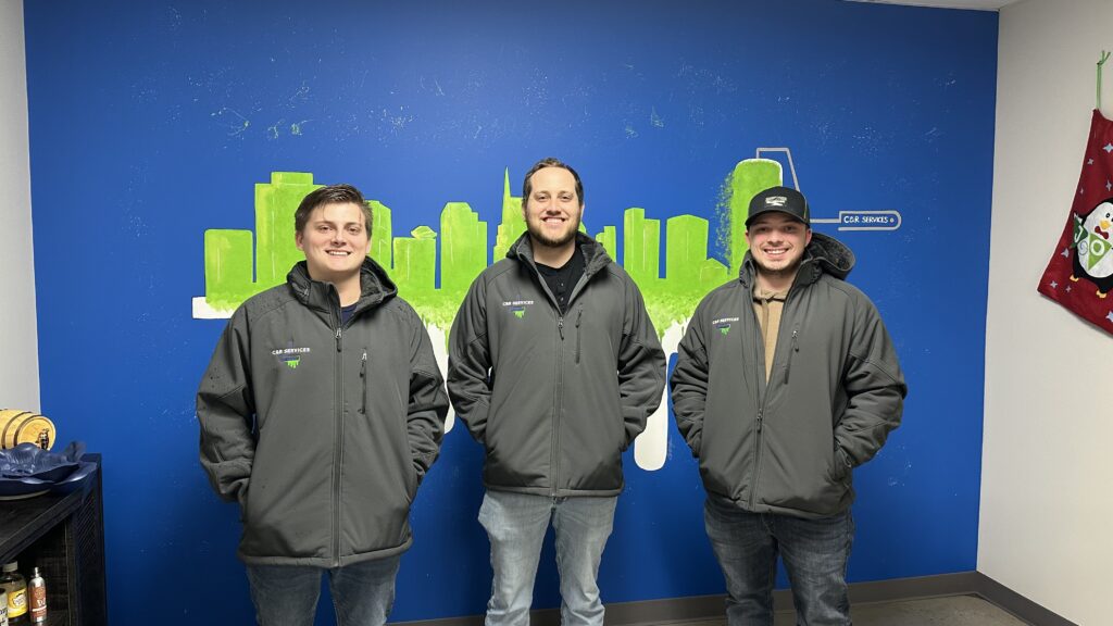 Three C&R Services team members wearing business branding jackets posing at their office.